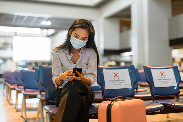 Person sitting cross-legged on an airport chair looking at a phone. A suitcase is in front of the image. A chair next to the person has a large, red X and read "Social Distancing."
