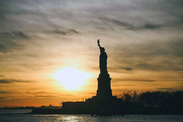 Statue of Liberty in shadow in New York Harbor as the sun sets behind