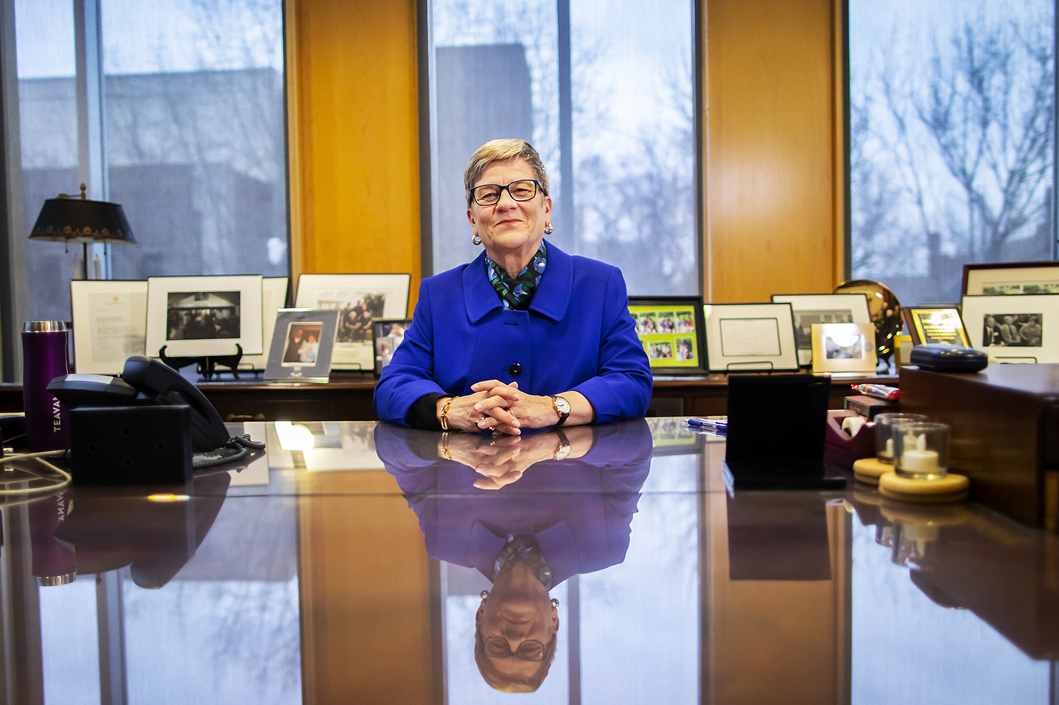 Person sitting at a large desk that includes a phone and some other items. Picture frames line a desk in the background.