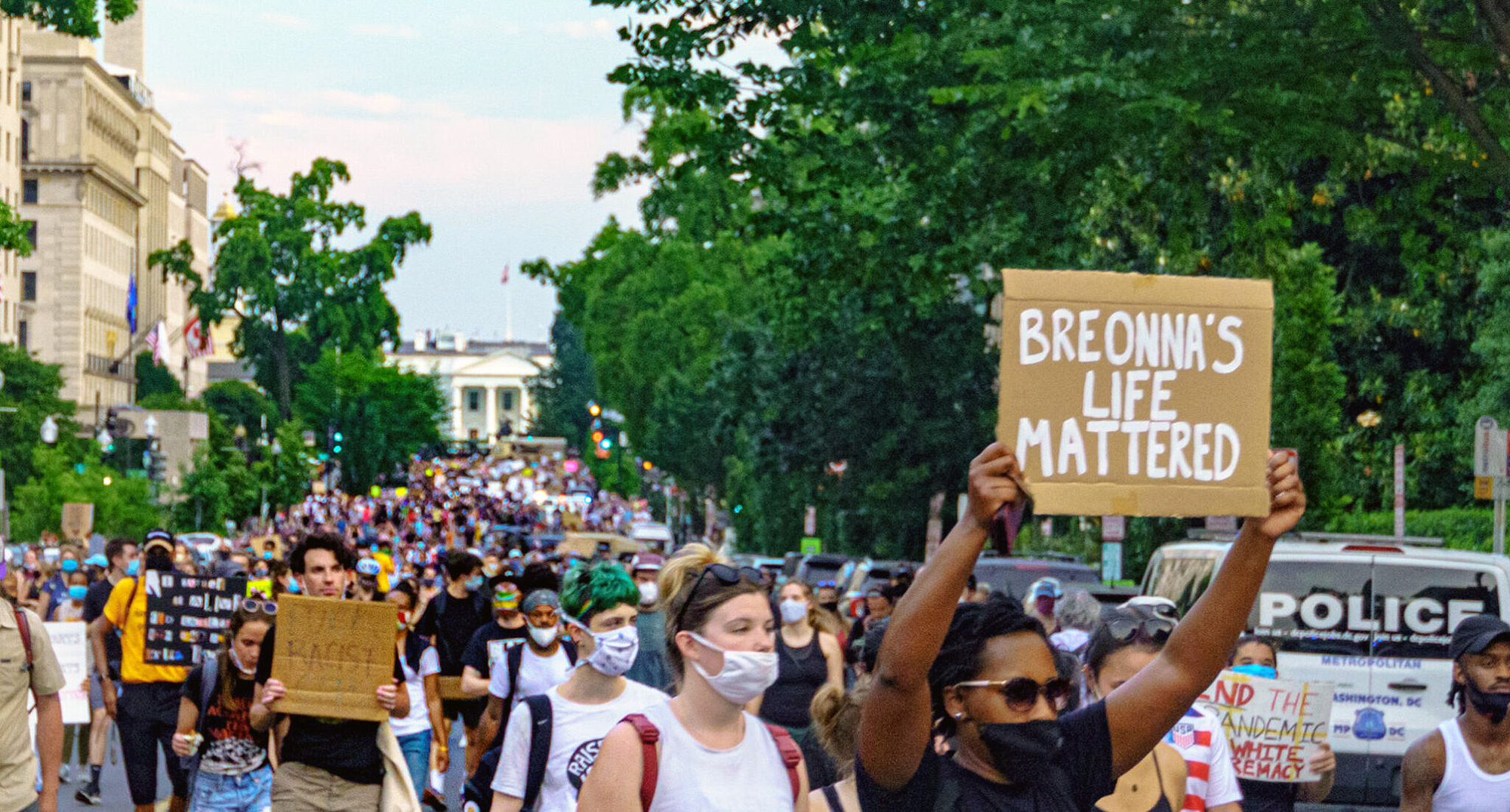 Large crowd wearing masks protesting in the streets of D.C., person in foreground holds a sign reading BREONNA’S LIFE MATTERED.