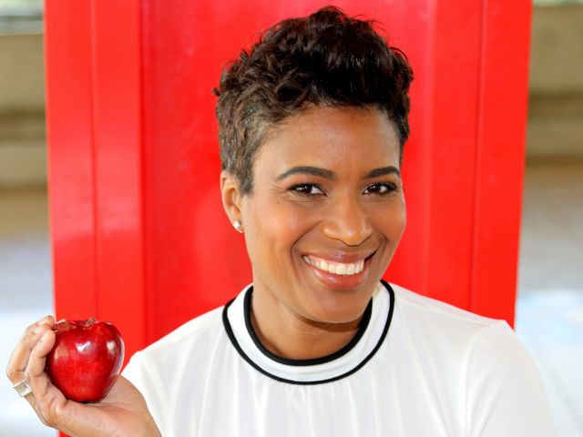 Smiling person holding an apple, standing in front of a red screen. 