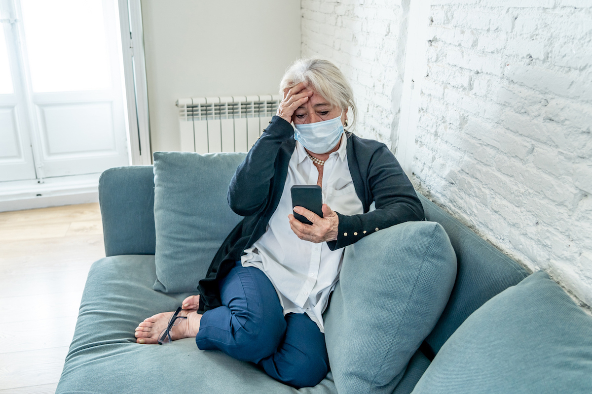 Distressed person wearing a mask sitting on a couch looking at a phone, hand on forehead.