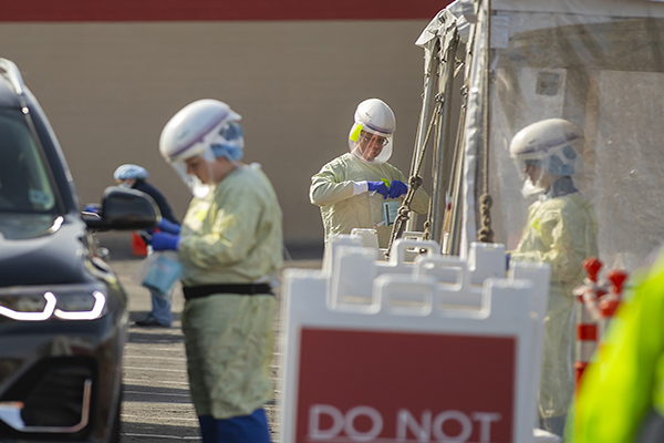 Three medical personnel in full PPE working at a drive-up COVID testing site.