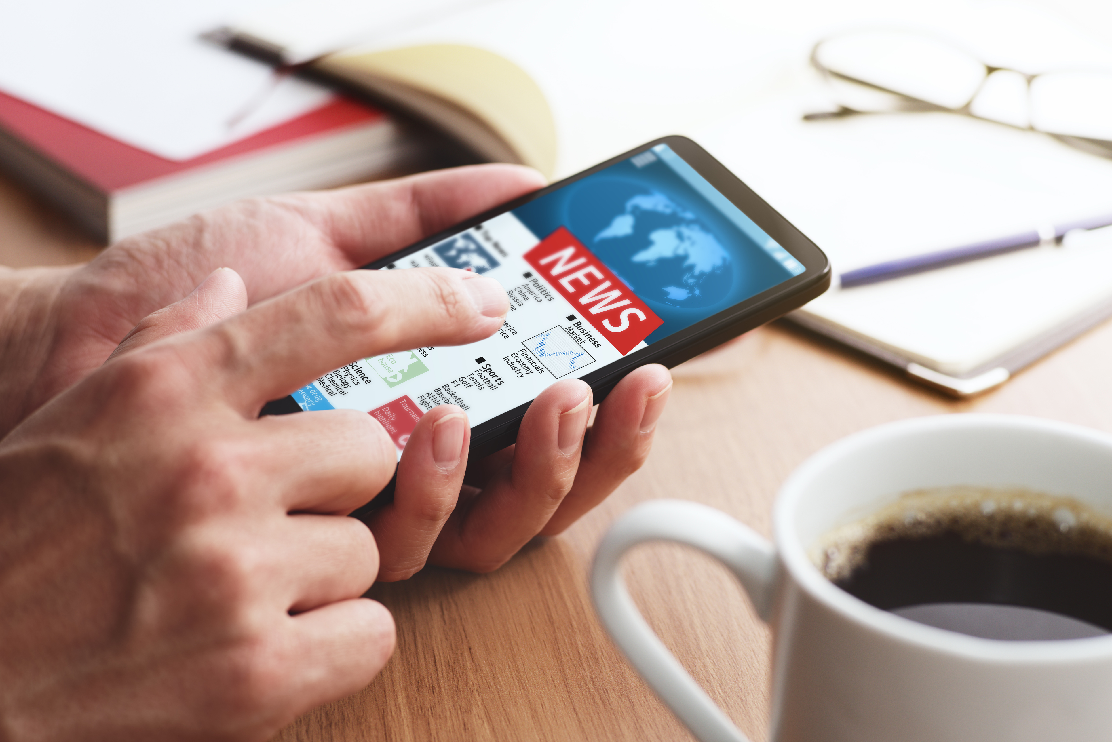 A close-up image of a hand holding a mobile phone, with the words "News," "Business," Politics" and "Sports" visible on the screen. Next to the phone is a cup of coffee. In the background are blurred open books.