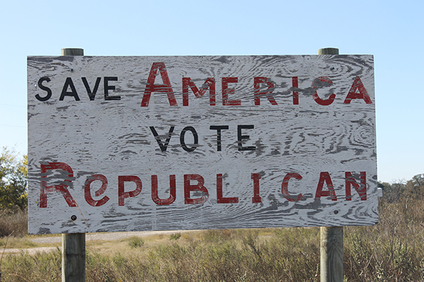 A faded wooden sign reading "save America vote Republican" stands amid scrubby plants on a Texas roadside.