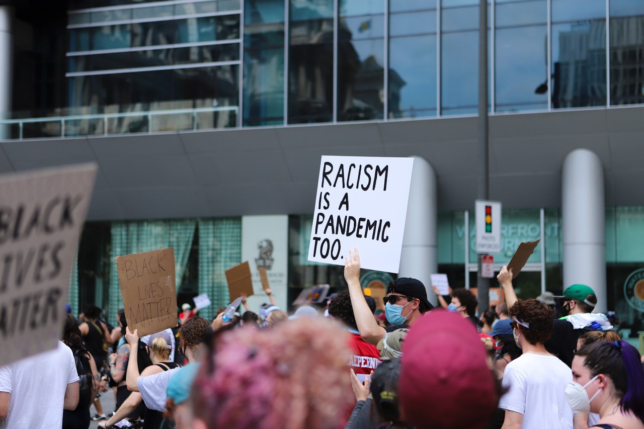 People marching during the pandemic, wearing face masks, one sign reads “Racism is a Pandemic Too”