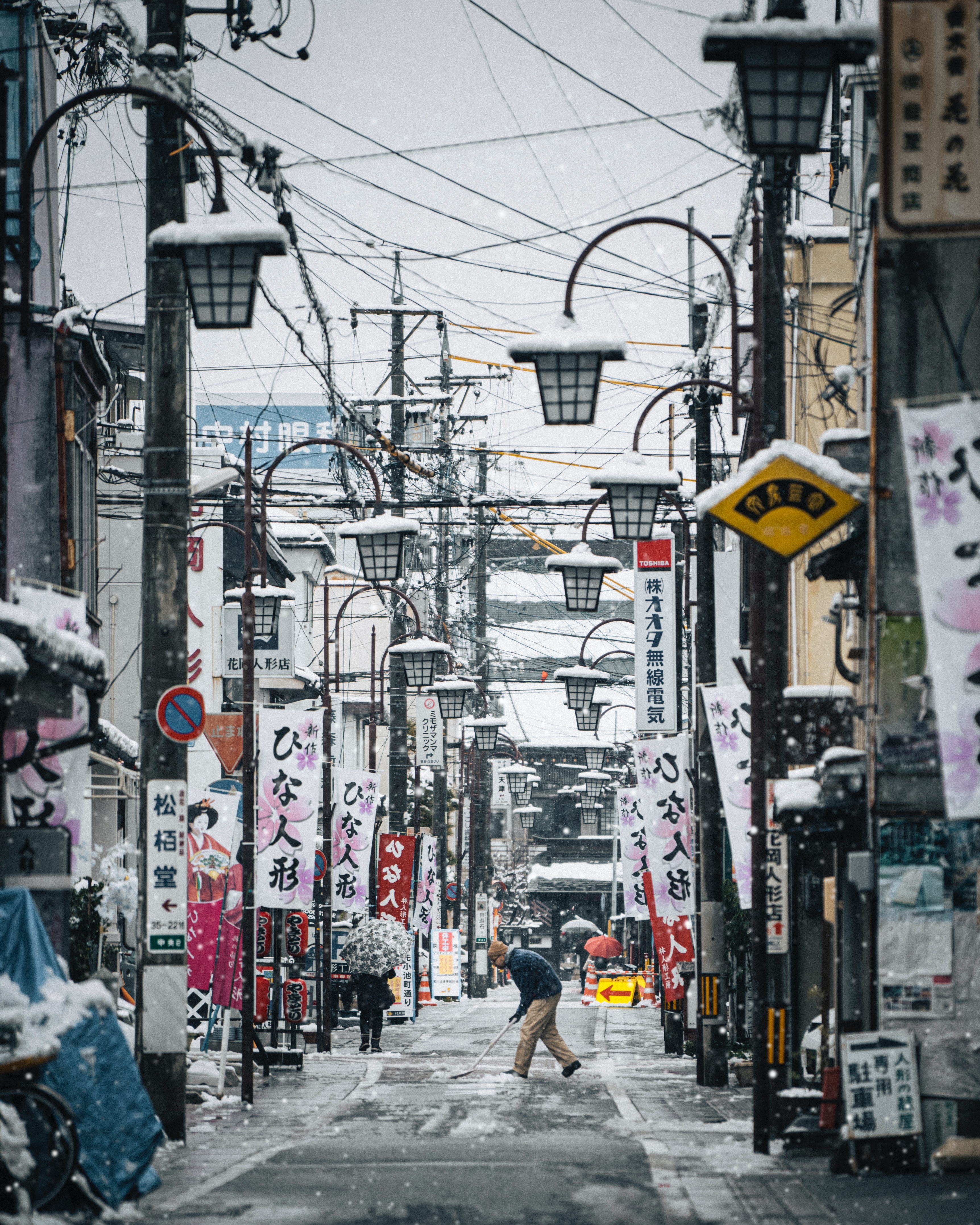 Man shovels snowy street, which is lined by lanterns and banners with Japanese characters