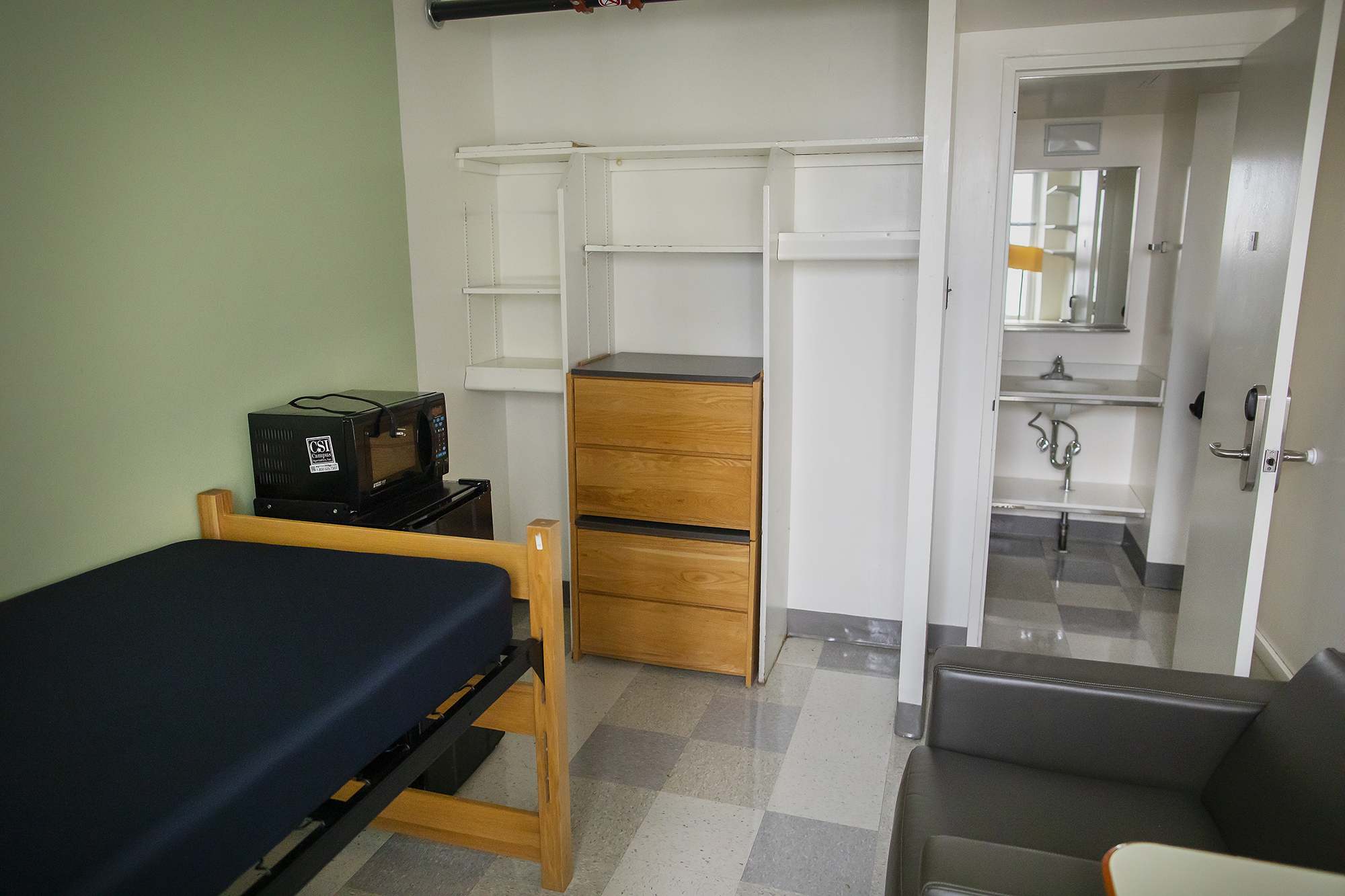 a dorm room with a bed, micro-fridge, dresser, chair