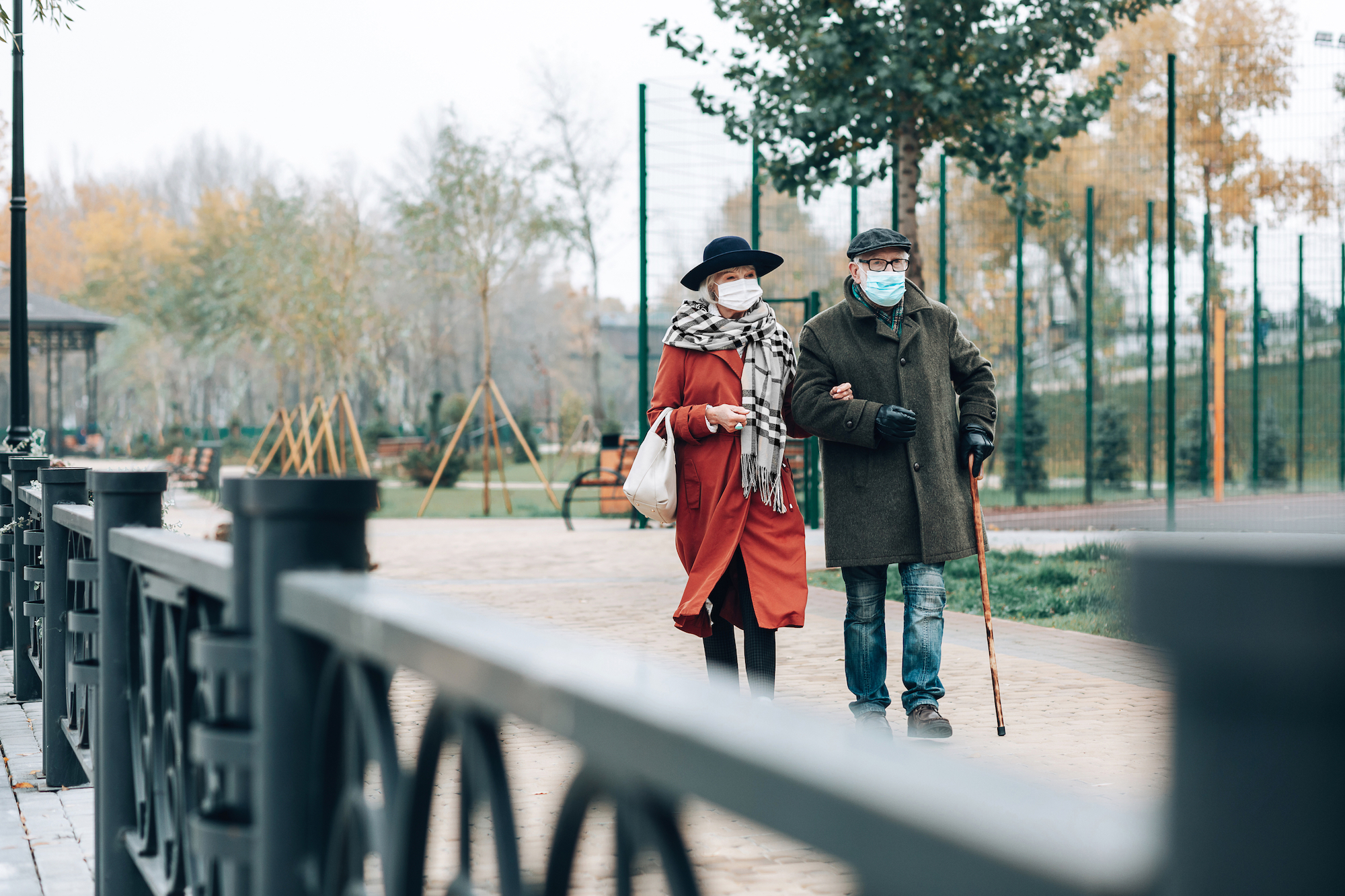 Two older adults walking outside, wearing cold-weather gear, walking arm in arm across a bridge, trees in the background.