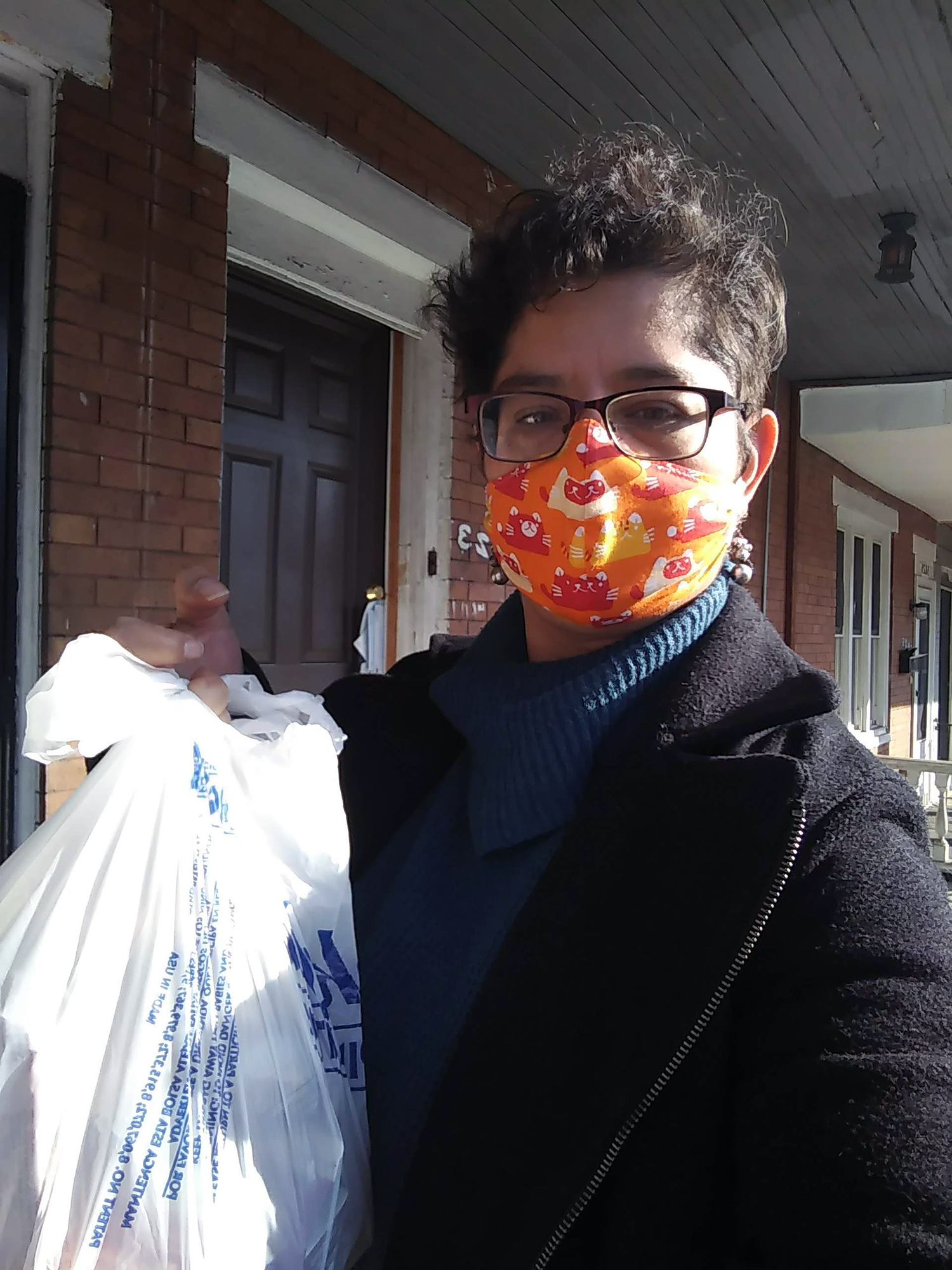 Sarvelia Peralta Duran wears a face mask and holds up a plastic shopping bag of groceries on the front porch of a house in Philadelphia.