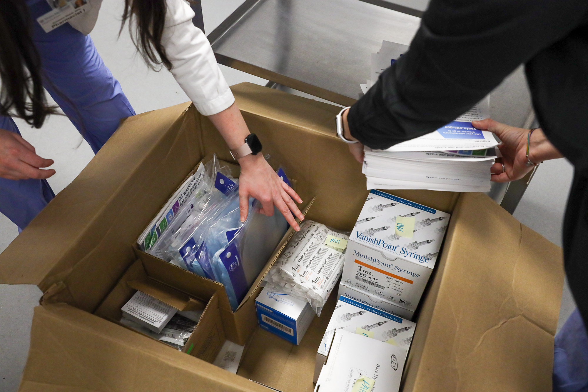 Two medical personnel unpack a large cardboard box containing syringes and medical equipment for vaccine distribution.