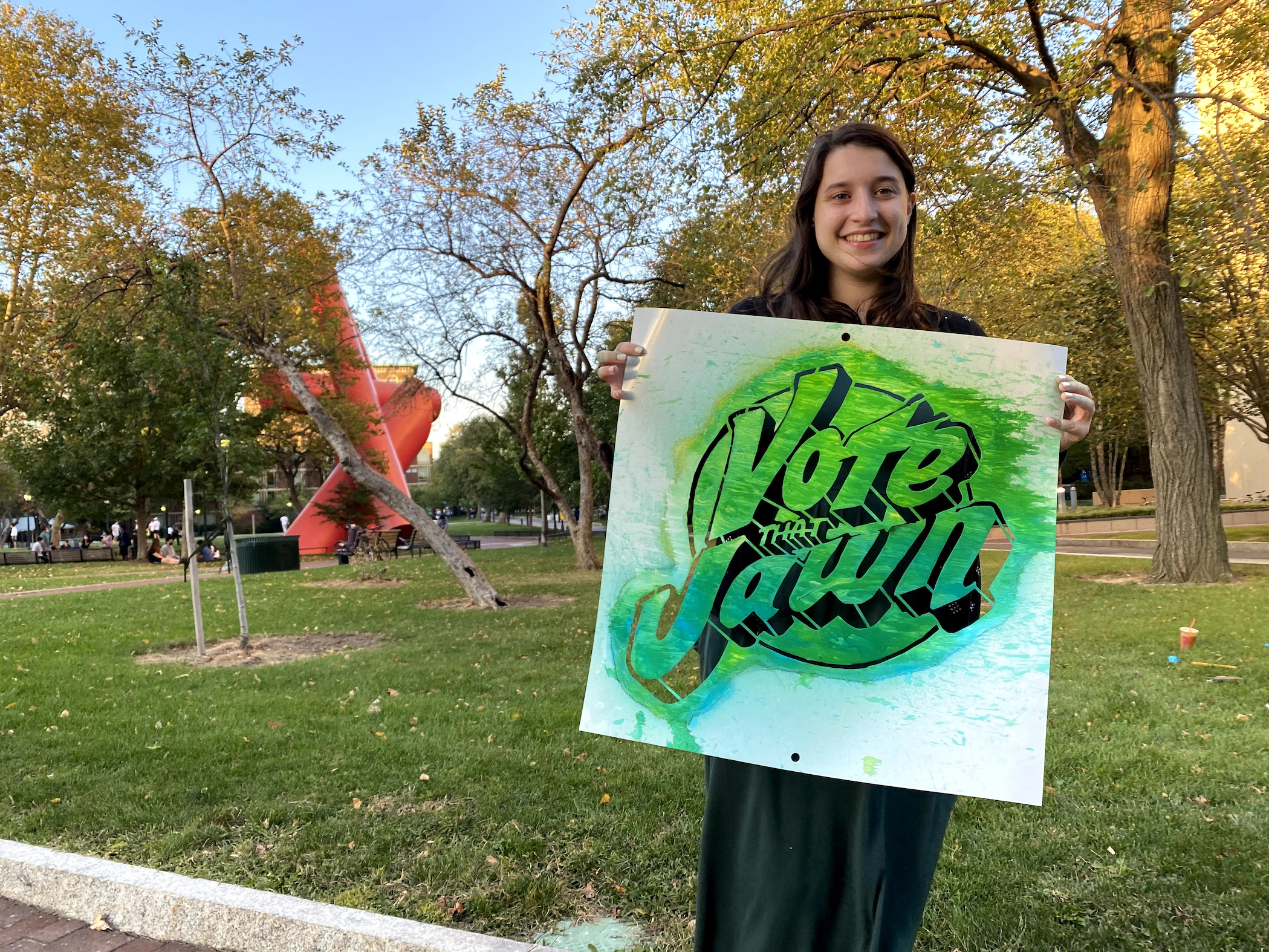 Student standing outside holding a sign reading Vote That Jawn with a towering sculpture behind her