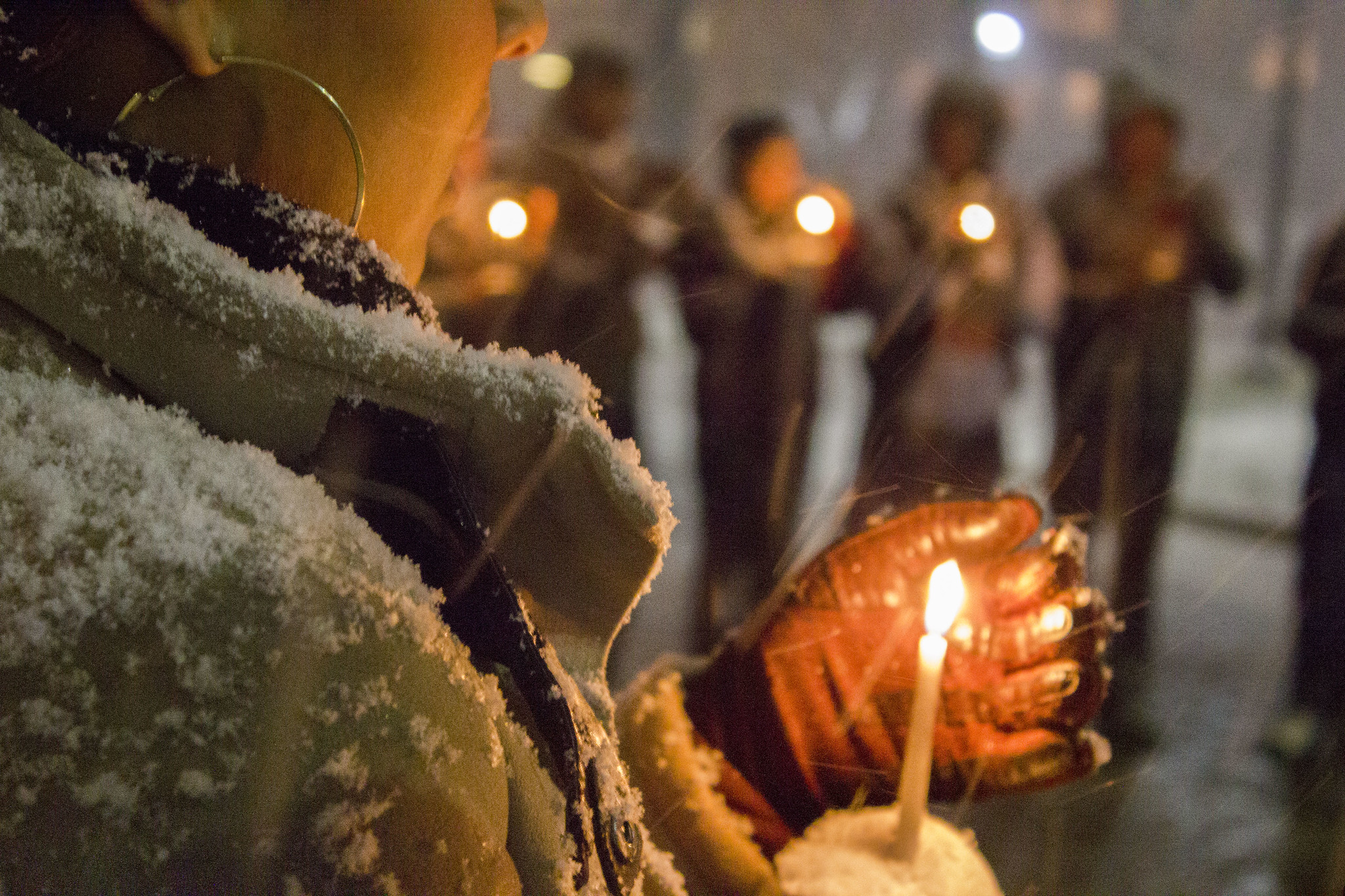 Woman cups a candle with a gloved hand. She is standing in a circle with other candle-holders in the snow.