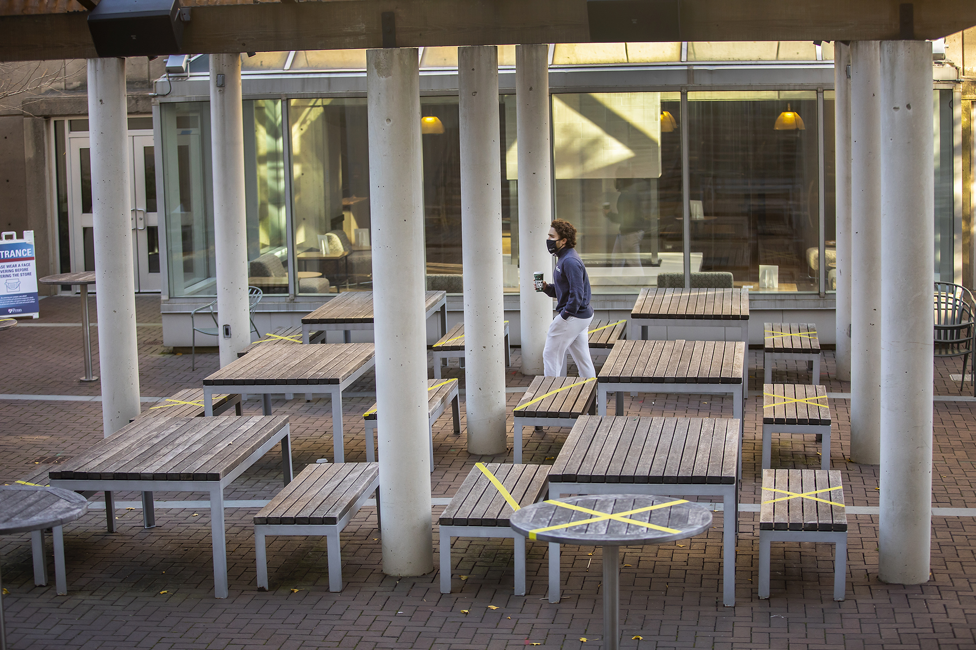 Empty tables at an outdoor dining area, some marked off from use for social distancing, with a student walking past wearing a mask