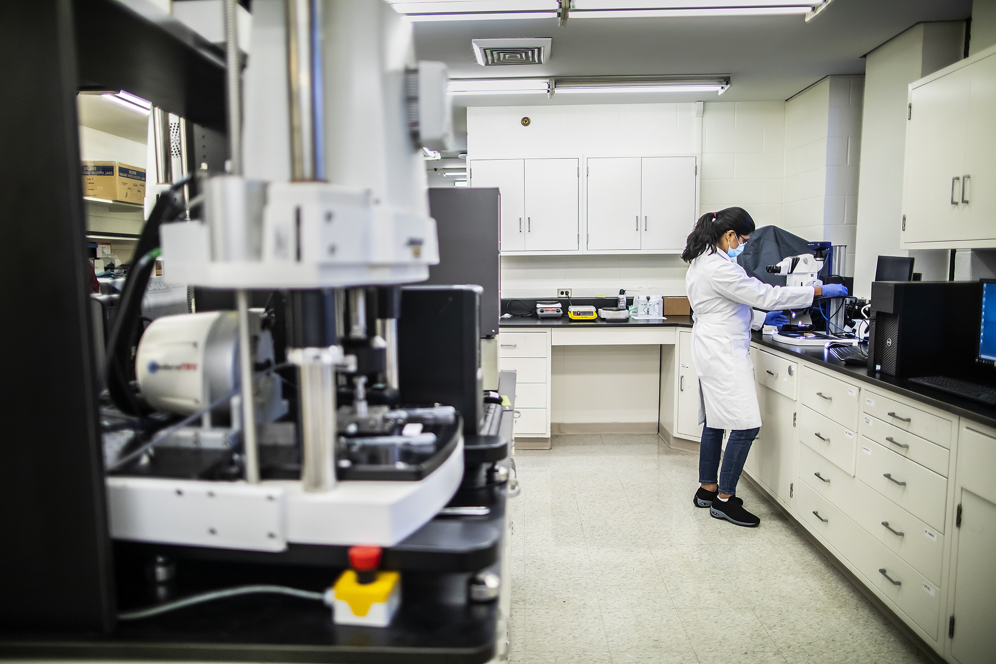 researcher in lab looks through microscope in background, framed by lab equipment in foreground