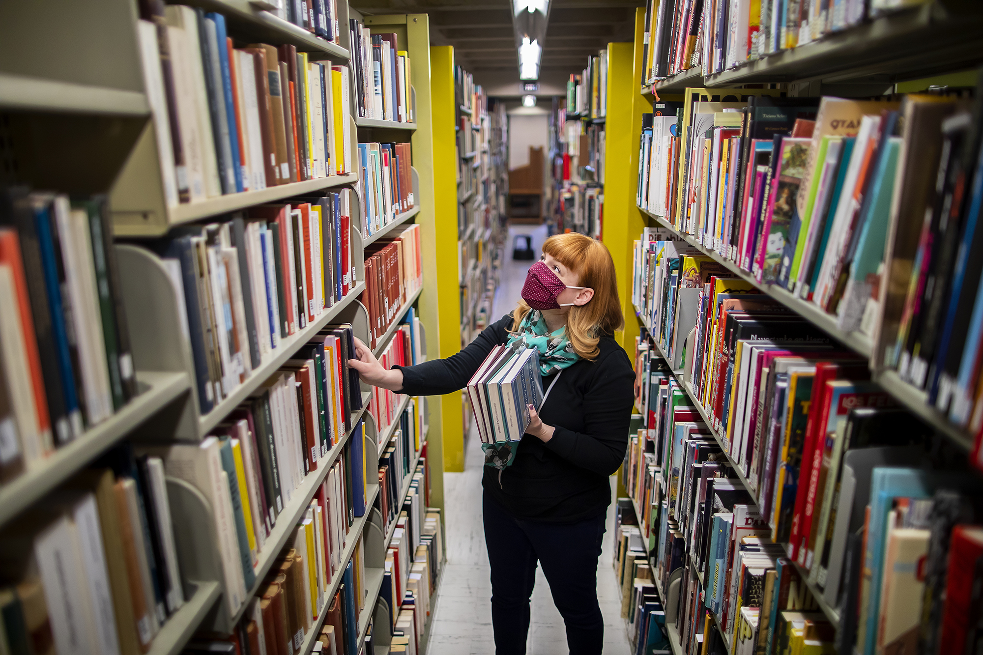 Person wearing a mask stands in the library stacks holding books.