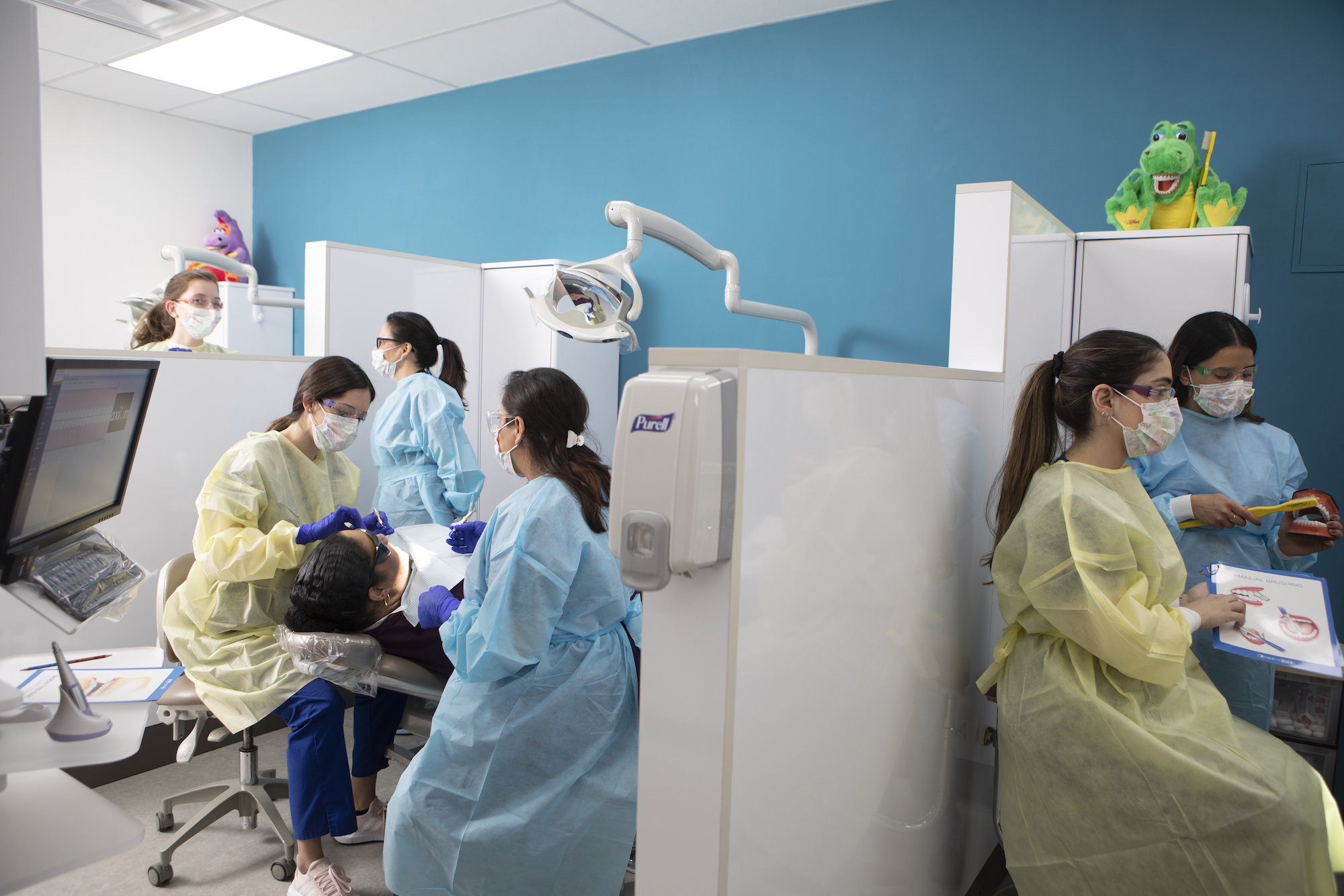 Dental professionals care for and educate patients in chairs while wearing gowns, masks, and gloves