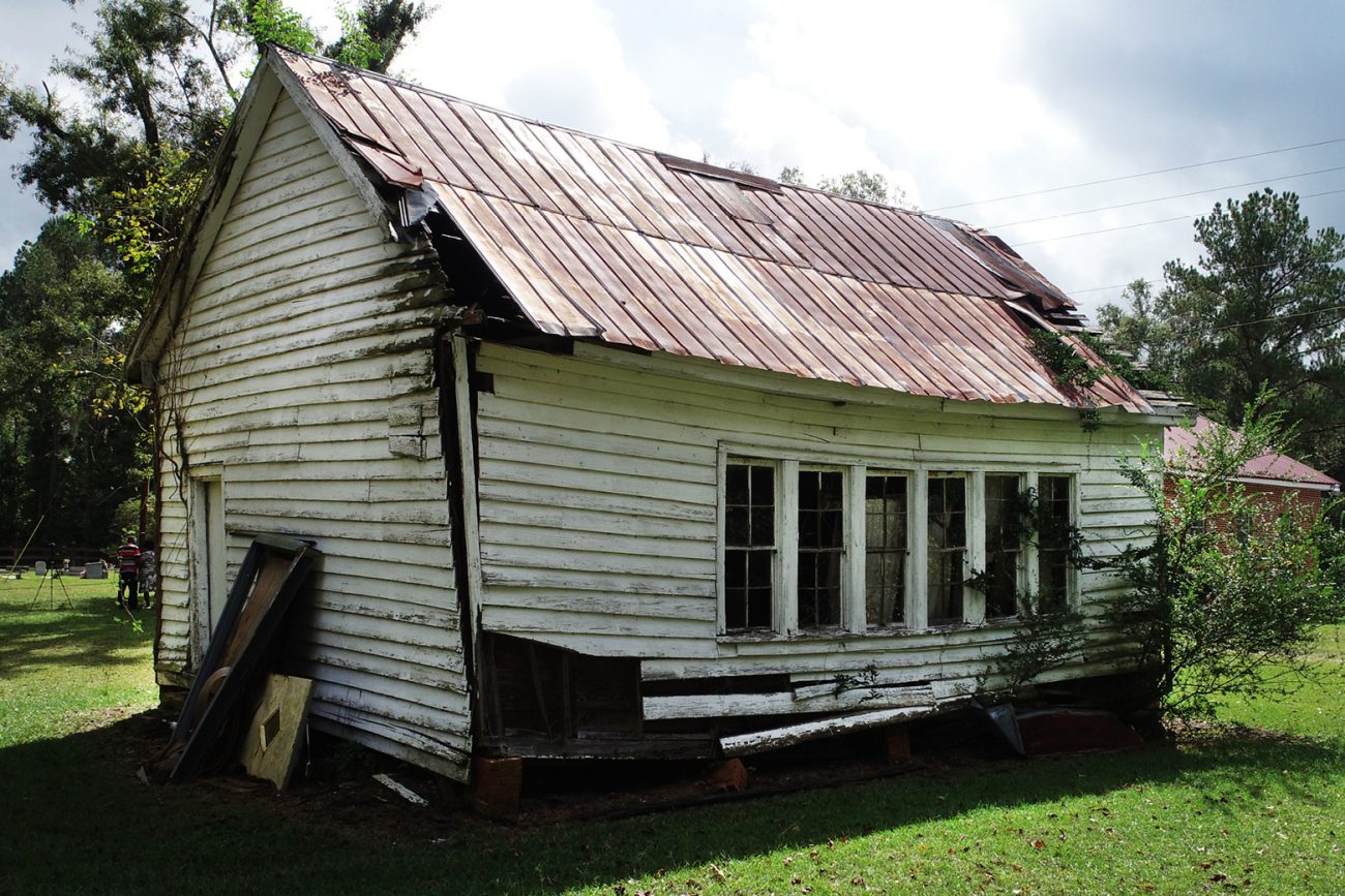 A rural one room schoolhouse with damage to its structure--boards coming loose, holes in the roof