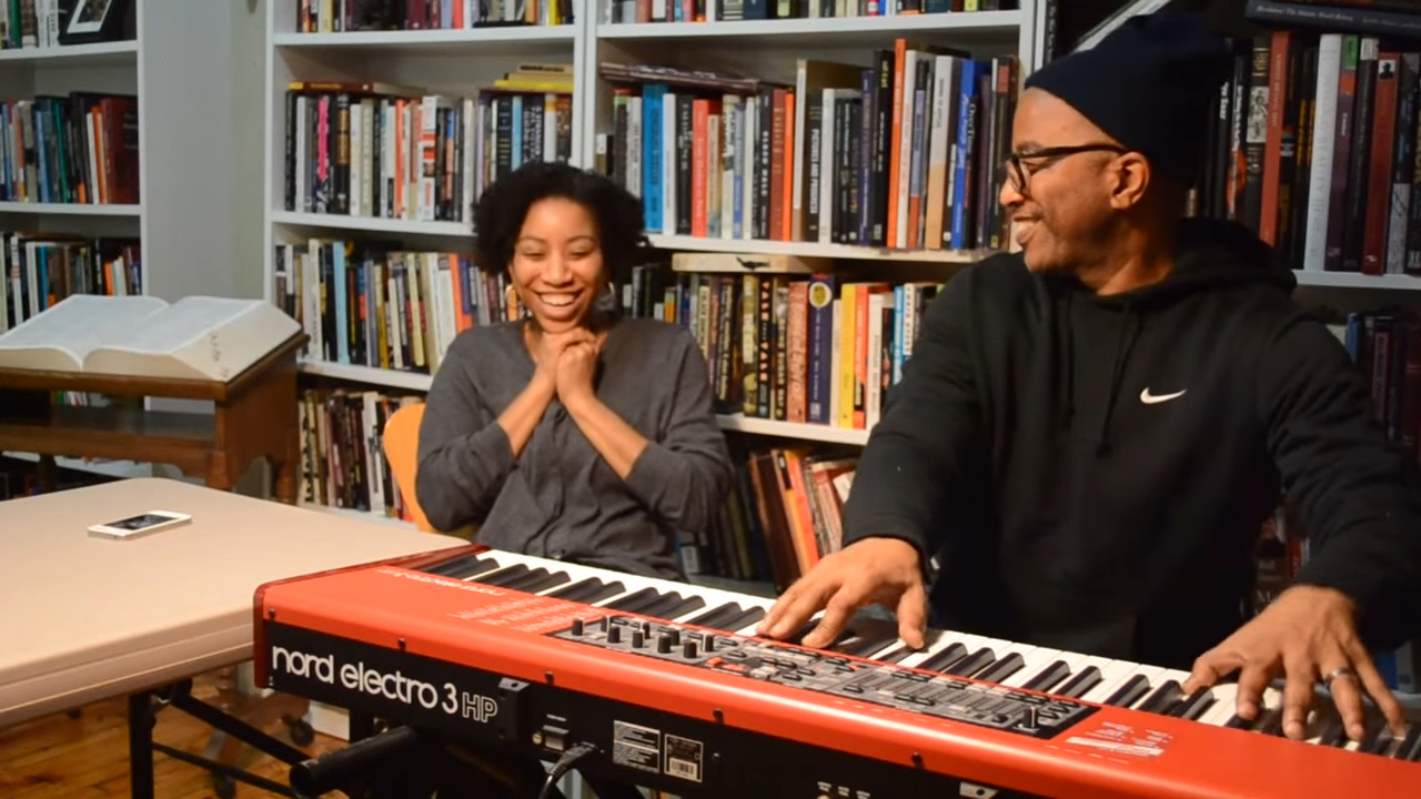 Father plays keyboard while his daughter smiles. Bookshelves in background. 