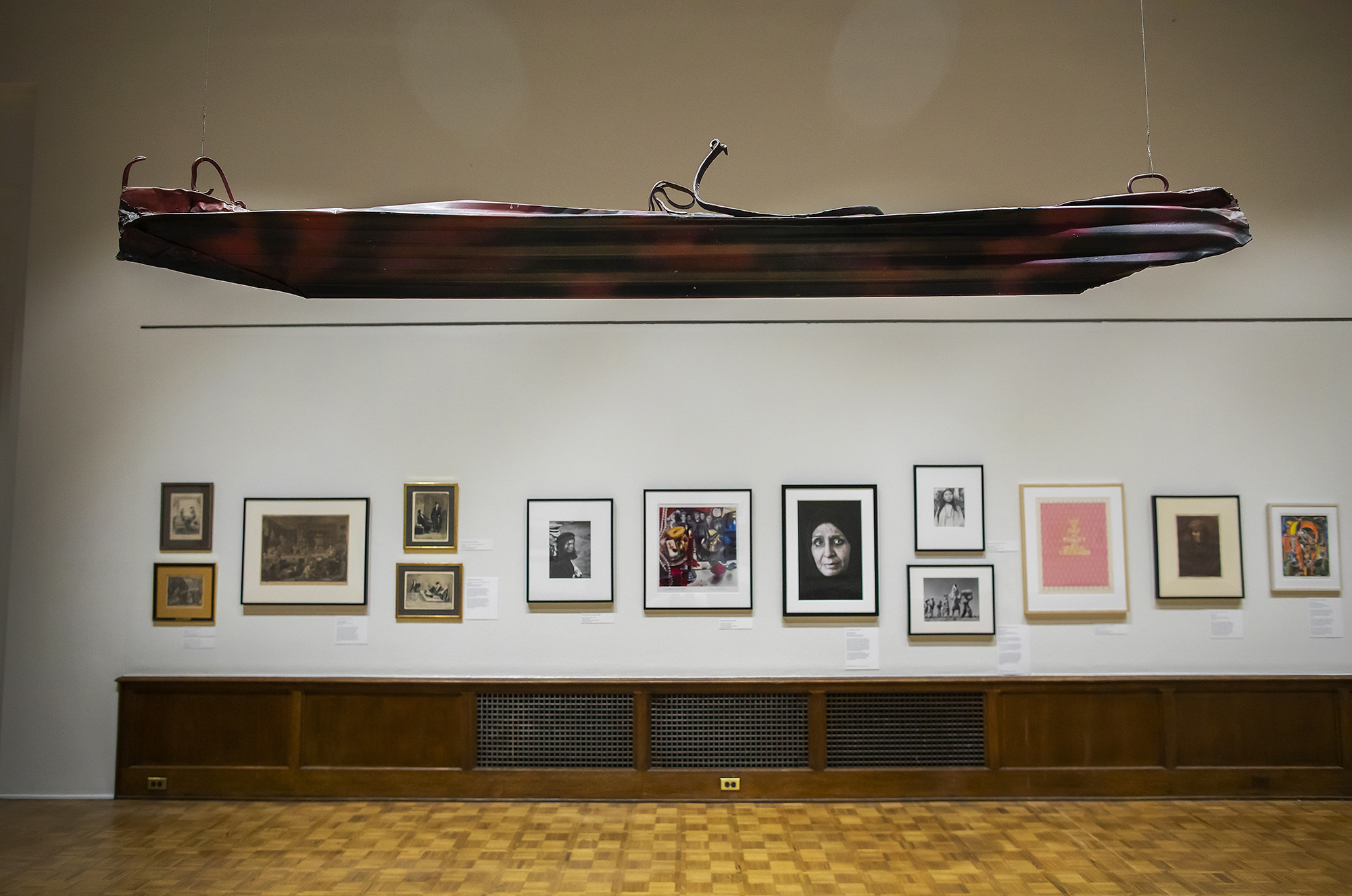 A metal kayak hangs from the ceiling in an art gallery with several artworks on the wall.