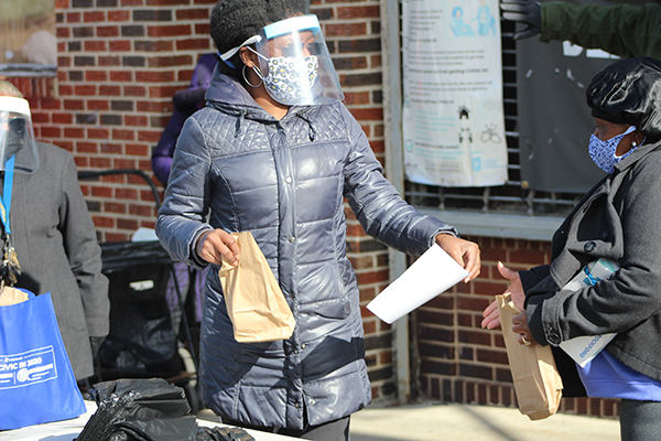Person standing outside holding a brown lunch bag to distribute wearing a face shield and mask outside a building in West Philadelphia