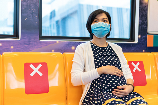 Pregnant person sits on a seat on public transit wearing a face mask.