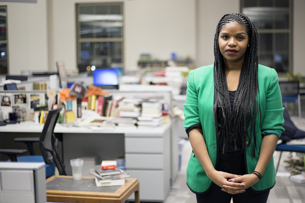 A woman with braids stands in an empty communal office space with books and computers on the desks