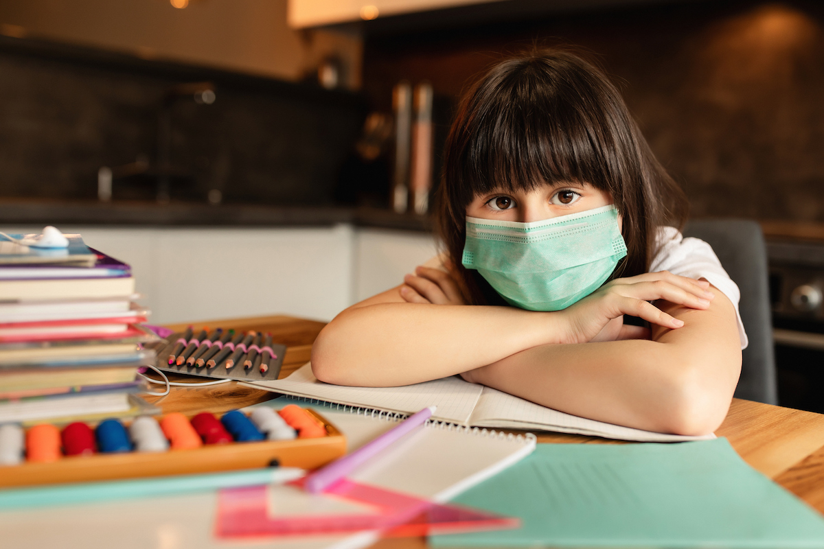 Young person sitting at a table, leaning chin on crossed arms, wearing a mask. There are books, colored pencils, an abacus, a notepad and more scattered around. 