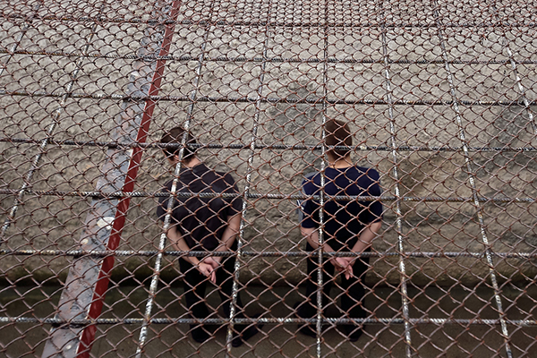 Two incarcerated people seen from above and behind with their hands handcuffed behind their backs outside in a prison yard.