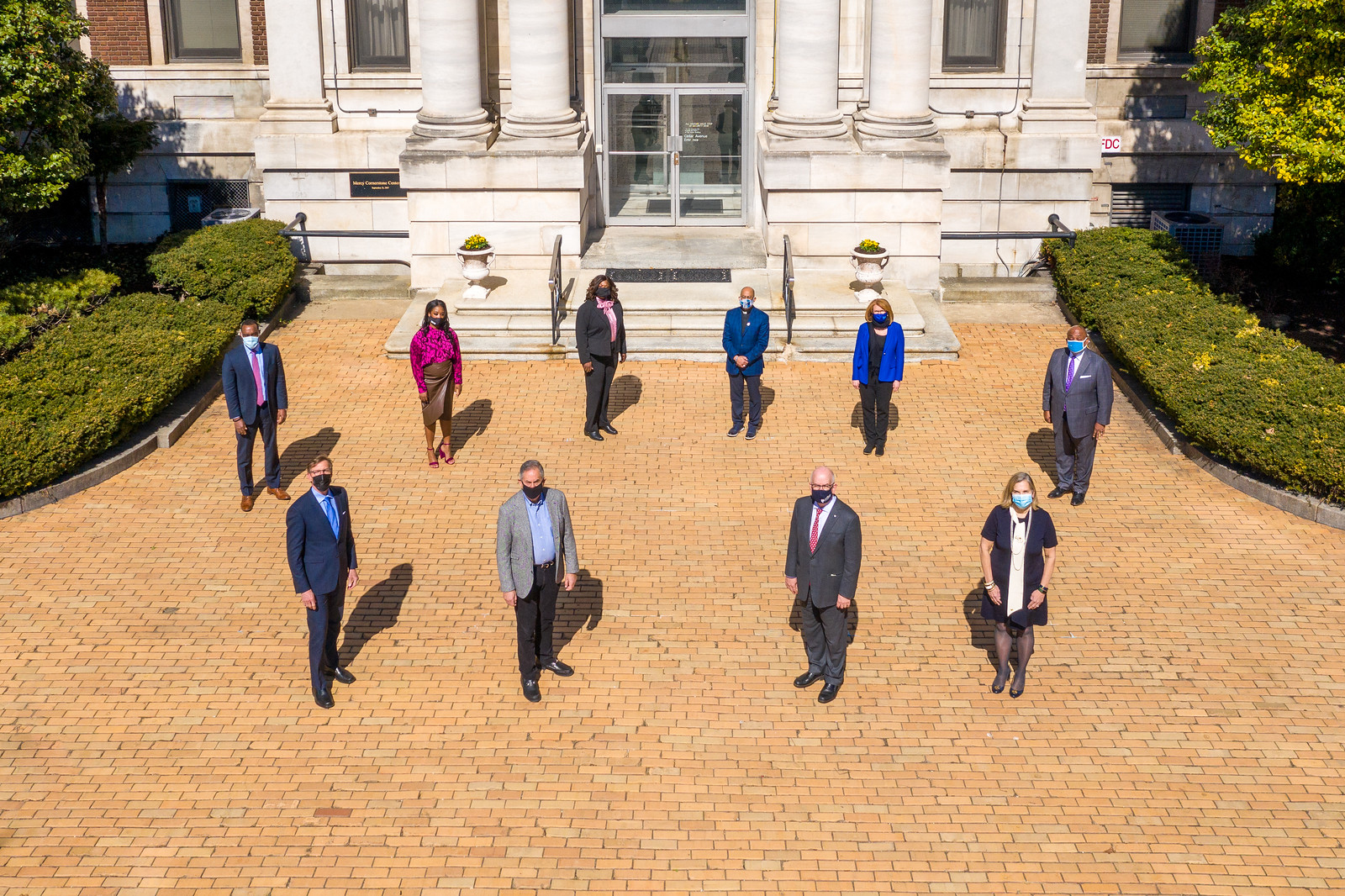 Aerial view of ten people wearing masks and social distancing in two rows on the front walkway of the Mercy Medical building. 