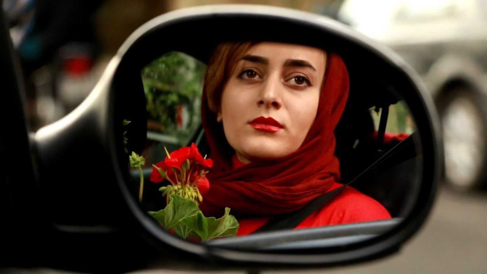 A woman with a red headscarf and red red jacket, and red lipstick holding a red geranium is seen in a reflection in a car's side mirror