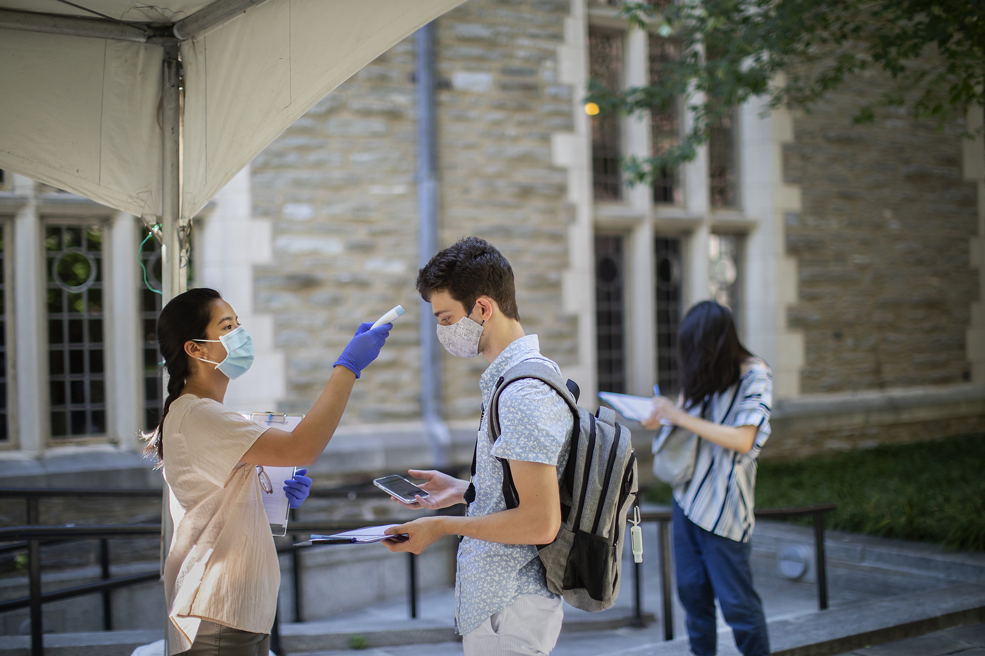 Student volunteer wearing a face mask scans the temperature of a masked Penn student outdoors on campus.