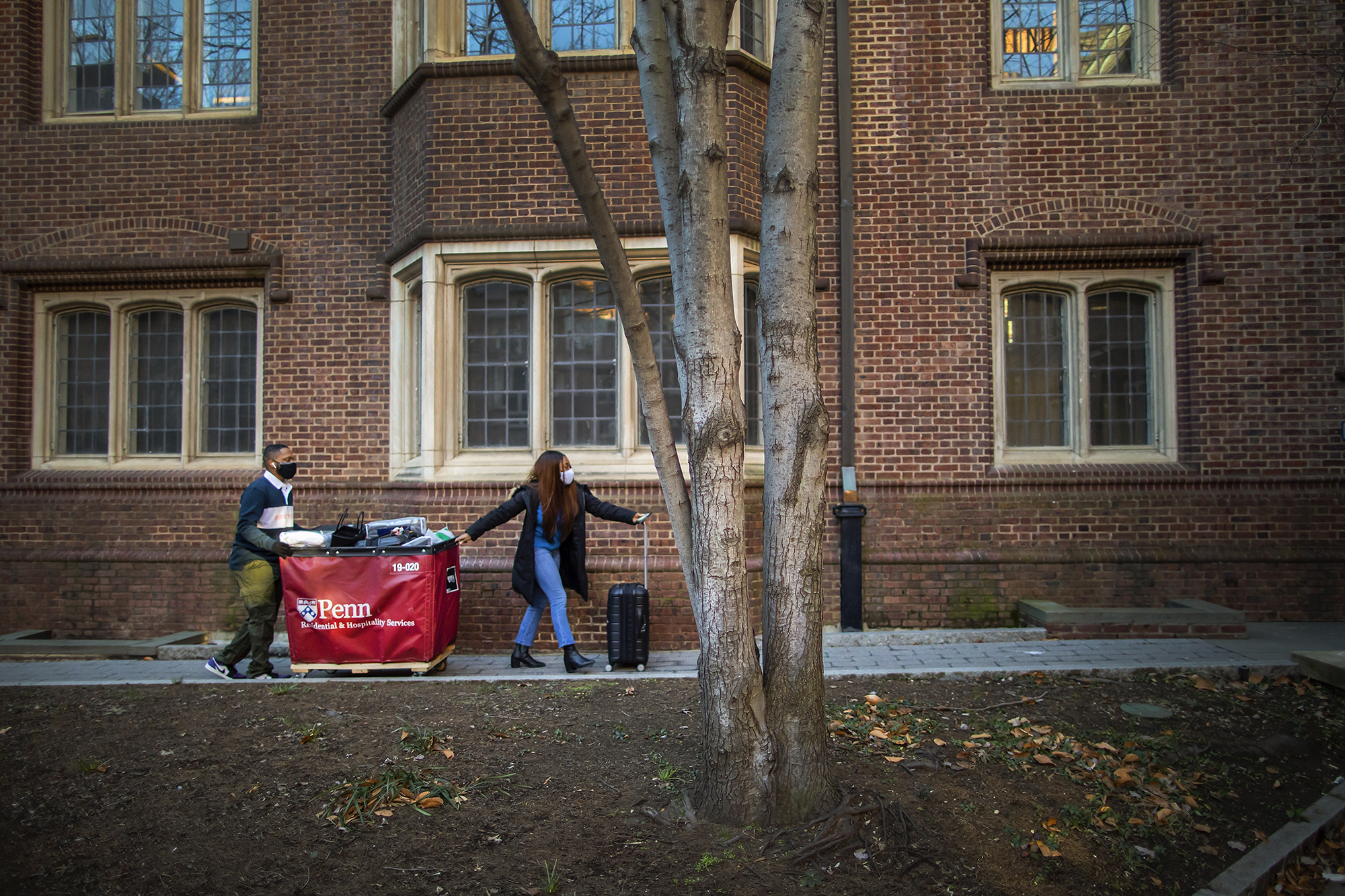 Two people wearing a face mask guide a Penn Move-In bin on wheels and suitcase outside a dorm room.