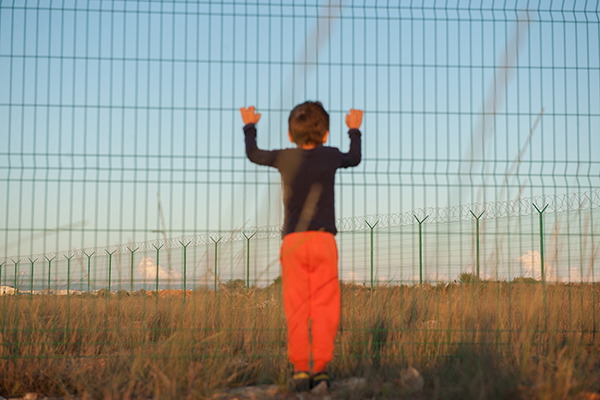 illegal immigration displaced persons camp concept of little boy in black and orange clothes holding fence with barbed wire in desert on state border