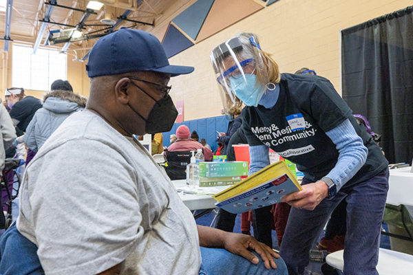 A community health care worker wearing a face shield and mask leans toward a seated community member in a face mask asking question prior to receiving a vaccine.