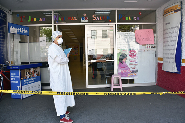 Nurse in a face mask and protective gear stands with a clipboard outside a hospital entrance that is roped off with police tape.