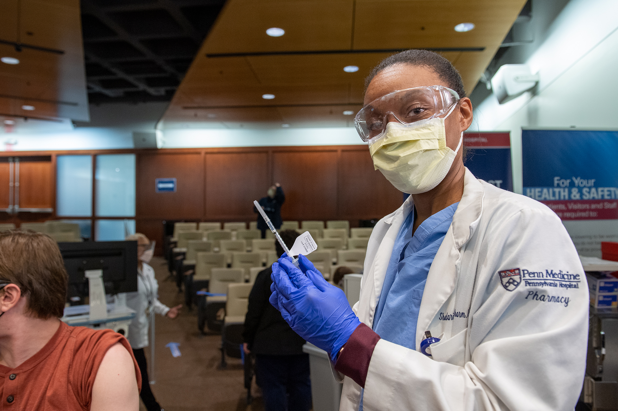 Medical professional with white coat and face PPE holds a syringe of COVID vaccine in a gloved hand, about to be injected in a person seated next to them with sleeve rolled up.