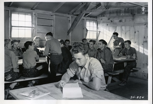 Man in foreground looks at pad of paper, behind him groups of young men sit at picnic tables inside a wooden structure with two windows on the back wall