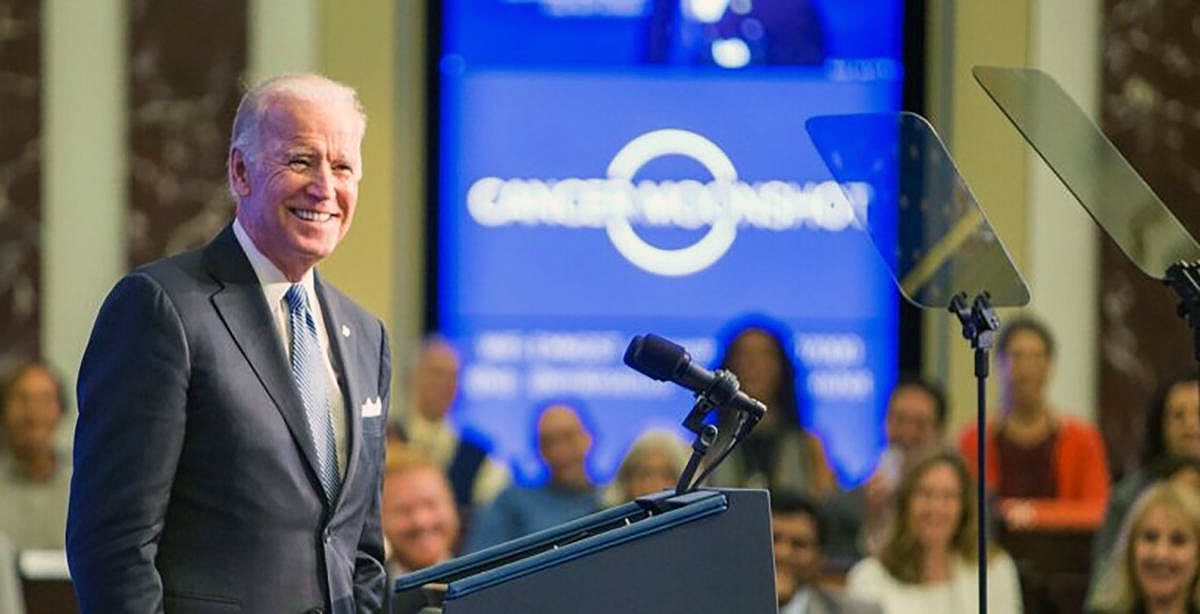 A man in a grey suit, light blue tie and white shirt smiles at a podium with a microphone as a crowd  that is blurred by the camera smiles behind him, beneath a blue display