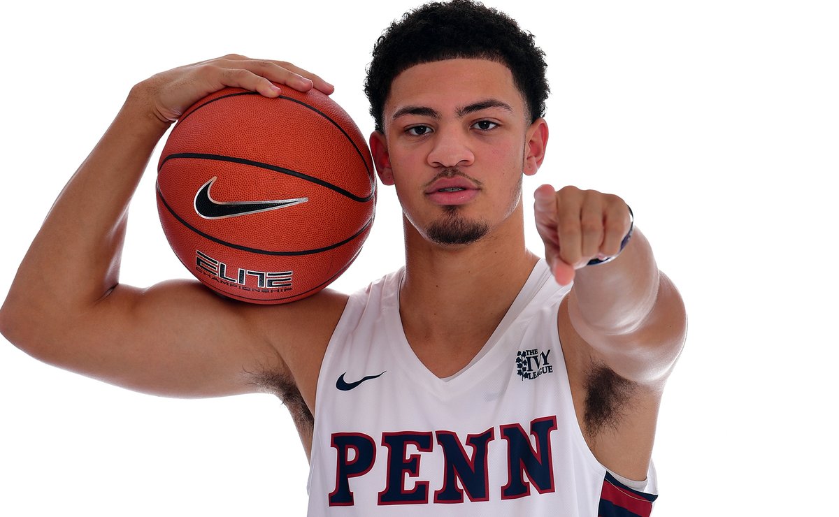 Jelani Wililiams, wearing a white Penn jersey, holds a ball on his shoulder and points at the camera.