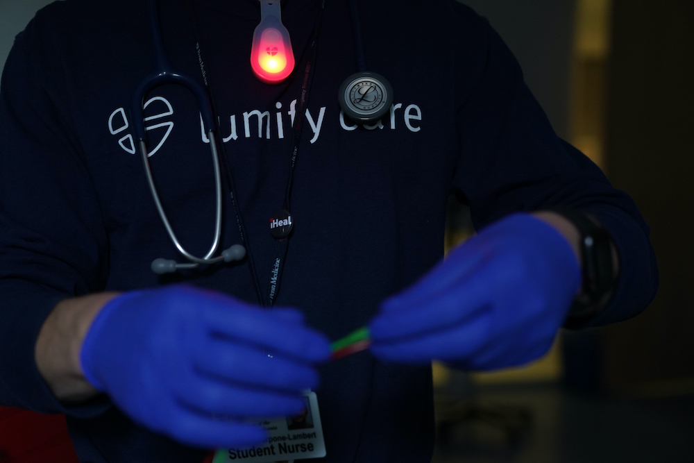 A torso wearing a stethoscope and a reddish clip-on light, in front of a shirt that reads "Lumify Care." In the front are blurry hands in latex gloves.