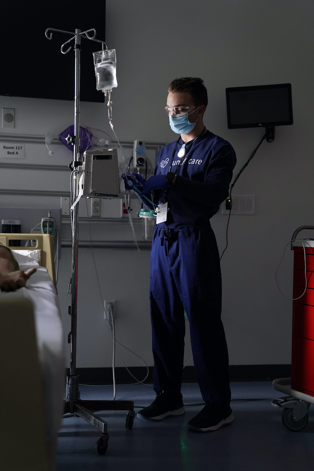 A nurse in scrubs and a face mask standing at a hospital bedside. On the shirt, below a clip-on wearable light read the words "Lumify Care."