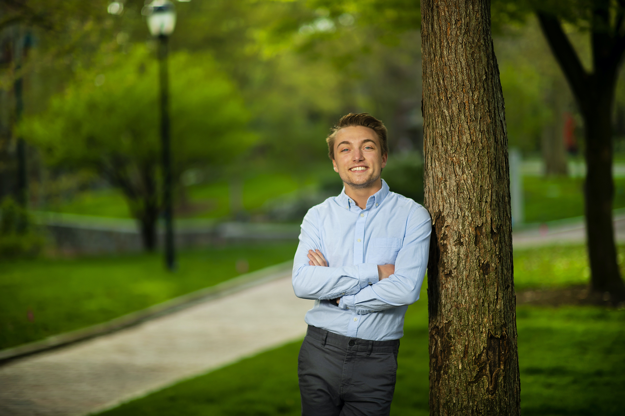 Person in a blue button-down shirt, arms crossed, leaning against a tree outside. In the background are blurred out bushes and trees, a path and a streetlight.