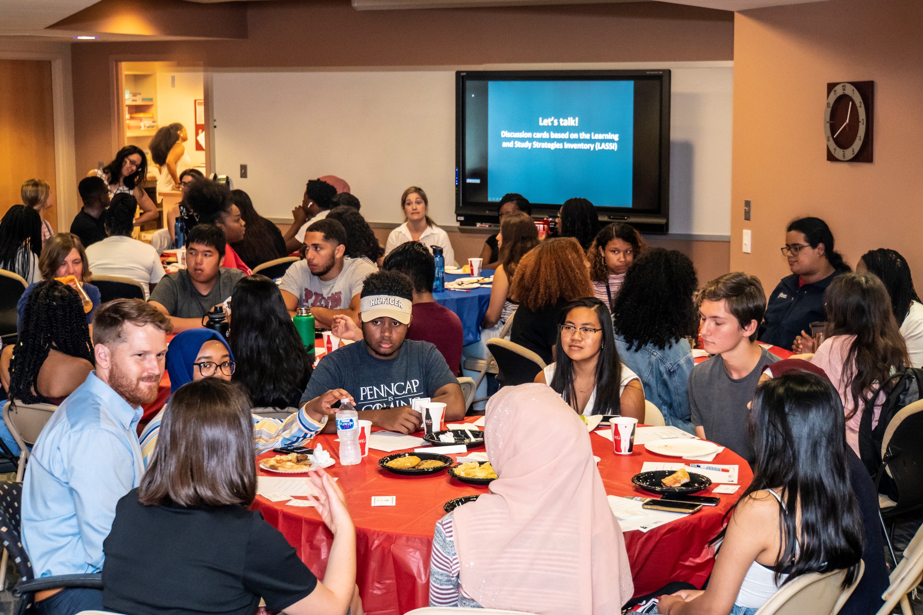 Students and staff sitting and eating at round tables. A screen in the back reads, "let's talk!"