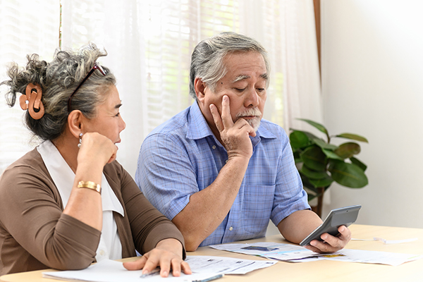 Two senior citizens look at a calculator while seated at a table covered in paperwork.