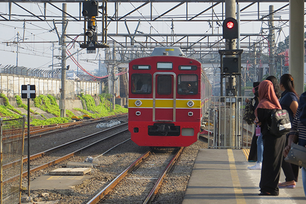 A red and yellow train approaches a platform as passengers wait on the right side, including one in a hijab.