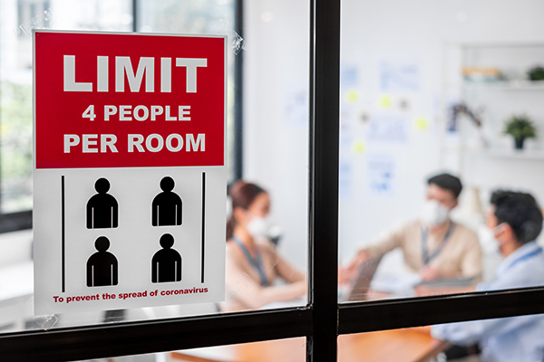 Sign on conference room window that reads LIMIT 4 PEOPLE PER ROOM, behind the window sit two masked employees.