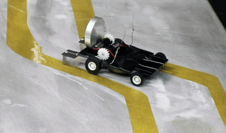 Film still of a small wheeled robot traveling a path between yellow tape.