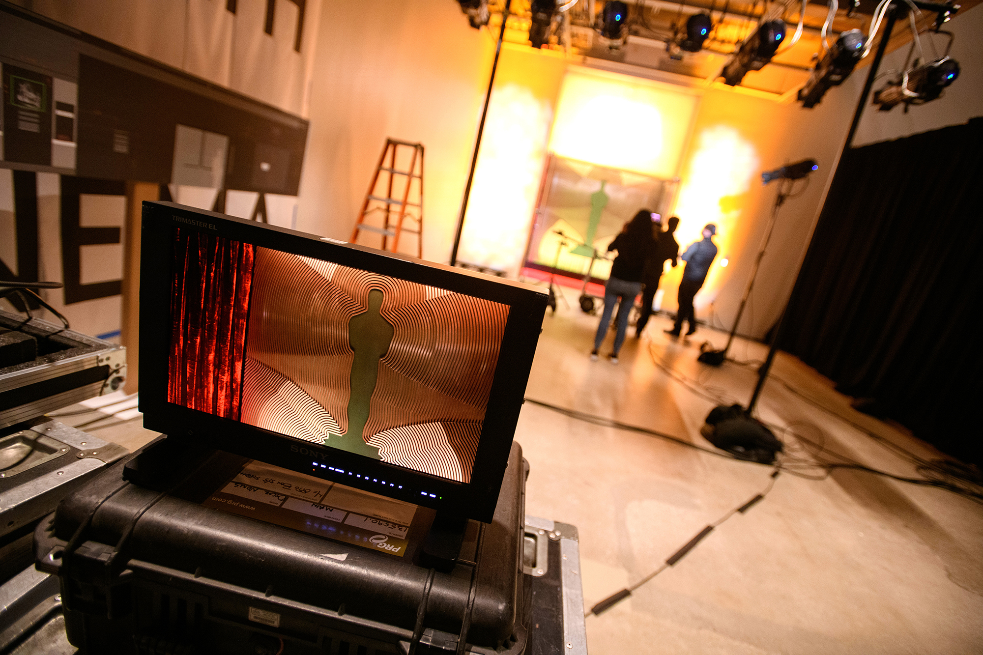 Video screen with shadow of the Oscars statue in foreground, three people in a photo studio taking photos of a screen on a wall illuminated with impact lights in background.
