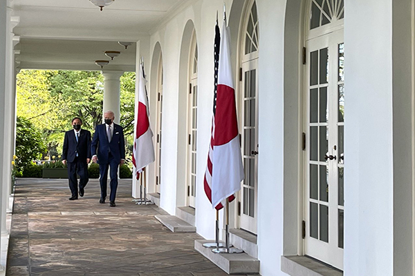 Two men in dark suits and dark face masks walk in the West Wing colonnade at the White House, past two sets of American and Japanese flags